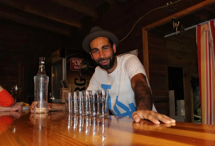 Man in a hat smiling behind a row of shot glasses at the bar